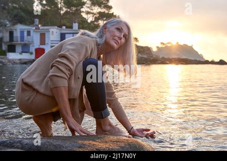 Grauhaarige Frau, die am Strand die Hand in Wasser auf Felsen taucht Stockfoto