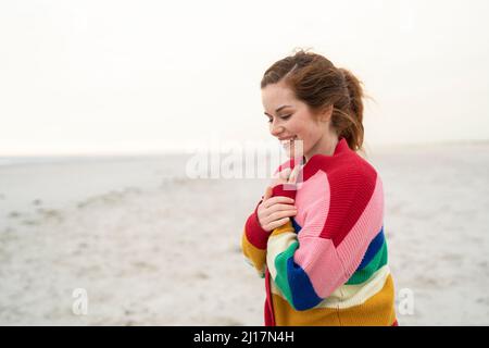 Fröhliche junge Frau, die einen mehrfarbigen Pullover am Strand trägt Stockfoto