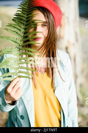 Lächelnde Frau, die im Wald das Gesicht mit Farnblättern bedeckt Stockfoto