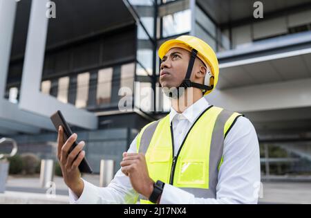 Mann mit Hardhut, der einen Tablet-PC hält und vor dem Gebäude steht Stockfoto