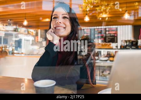 Eine durchdachte Geschäftsfrau mit der Hand am Kinn, die durch ein Glas Café gesehen wird Stockfoto