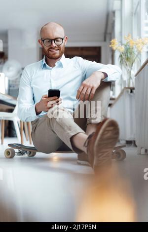 Lächelnder junger Geschäftsmann, der im Büro mit dem Mobiltelefon auf dem Skateboard sitzt Stockfoto