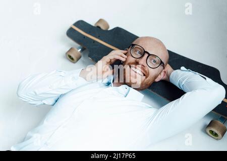 Glücklicher Geschäftsmann im Gespräch auf dem Handy liegend auf dem Skateboard im Büro Stockfoto