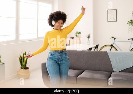 Junge Frau mit geschlossenen Augen hört Musik über kabellose Kopfhörer, die im Wohnzimmer tanzen Stockfoto