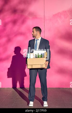 Ein Geschäftsmann trägt eine Box mit persönlichen Gegenständen vor einer rosa Wand Stockfoto