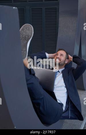 Lächelnder Geschäftsmann, der mit einem Laptop auf einem architektonischen Merkmal lügt Stockfoto