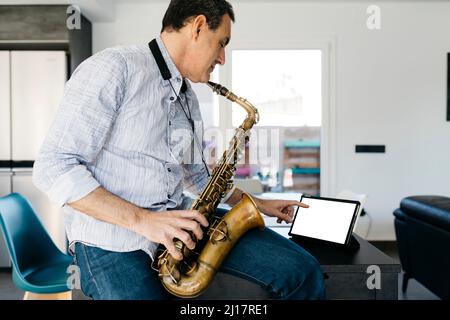 Musiker mit Saxophon mit Tablet-PC, der zu Hause auf dem Tisch sitzt+ Stockfoto