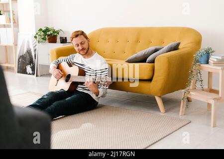 Junger Mann mit roten Haaren, der Gitarre trainiert, sitzt zu Hause vor dem Sofa Stockfoto