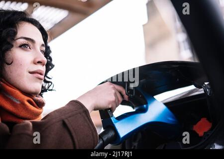 Junge Frau mit Ladegerät für Elektroautos an der Ladestation Stockfoto