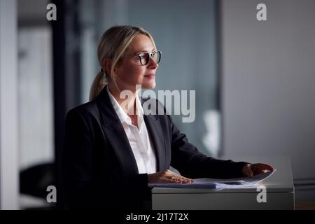 Blonde Geschäftsfrau mit Brille, die mit Dokumenten am Schreibtisch im Büro steht Stockfoto