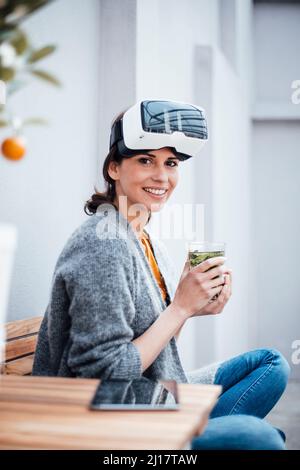 Lächelnde Geschäftsfrau im Virtual-Reality-Simulator mit Trinkglas auf einem Stuhl Stockfoto