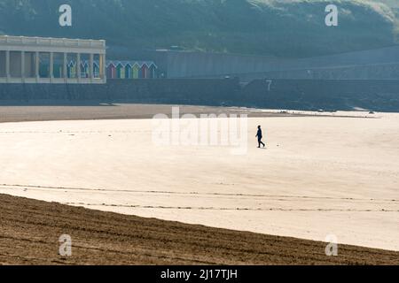 Eine einsame Person geht am frühen Morgen an einem sonnigen Sommertag über den Sandstrand von Whitmore Bay, Barry Island. Stockfoto