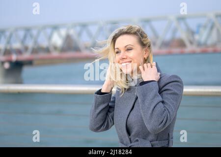Attraktive Frau, die an einem windigen Tag an einer Uferpromenade steht und ihre langen blonden Haare mit einem Lächeln anklammert Stockfoto
