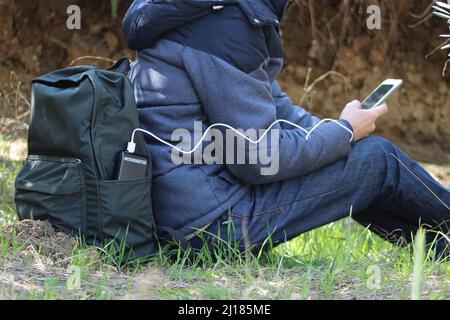 Der Mensch hält ein Smartphone in den Händen und lädt es vor dem Hintergrund eines Naturschauers mit einer schwarzen Powerbank auf. Tragbares Reiseladegerät. Stockfoto