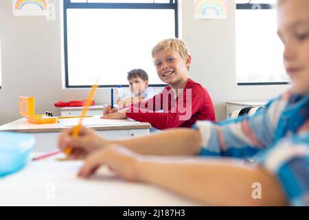 Porträt eines lächelnden kaukasischen Grundschuljungen mit männlichen Klassenkameraden, die im Klassenzimmer am Schreibtisch sitzen Stockfoto