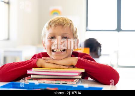 Porträt eines lächelnden kaukasischen Grundschuljungen, der sich auf gestapelte Bücher am Schreibtisch im Klassenzimmer stützt Stockfoto