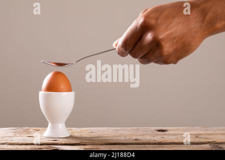 Beschnittene Hand hält Löffel über gekochtes braunes Ei in Tasse auf dem Tisch Stockfoto