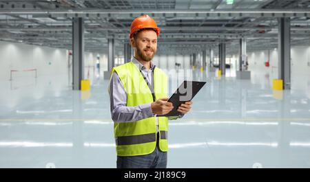 Ingenieur, der eine reflektierende Weste und einen Helm trägt und eine Zwischenablage hält und in einem leeren Lager auf die Kamera schaut Stockfoto