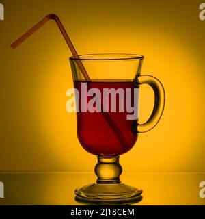Tee, Cocktail, Fruchtgetränk in einer Glastasse mit Trinkhalm auf dem Tisch. Rotes Getränk in einem Becher auf einem schönen, leuchtend gelben Hintergrund. Summe Stockfoto