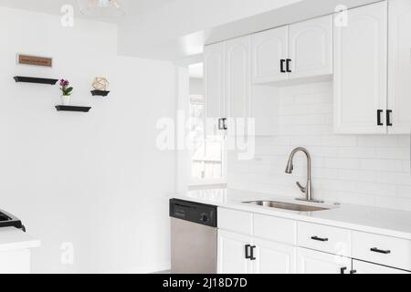 Eine helle Küche mit weißen Schränken, unterirdische Fliesenrücksplash, schwarze Hardware und ein Waschbecken aus Edelstahl. Stockfoto