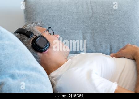 Mann, der auf dem Sofa liegt und schläft, während er mit Kopfhörern Musik hört Stockfoto