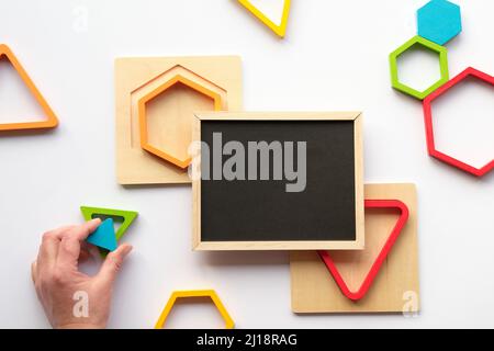 Geometrische Holzfiguren und Tafel. Genestete Holz-Dreiecke und Sechsecke in Regenbogenfarben. Türkisfarbenes Dreieck in menschlicher Hand. Stockfoto