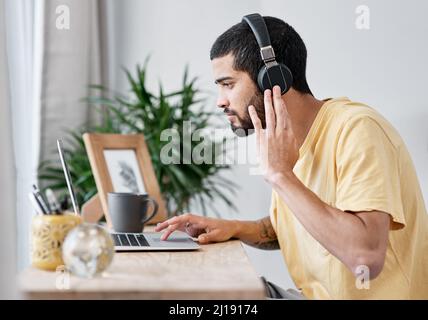 Die besten Kopfhörer mit Geräuschminimierung in der Branche. Aufnahme eines jungen Mannes, der einen Laptop und Kopfhörer benutzt, während er von zu Hause aus arbeitet. Stockfoto