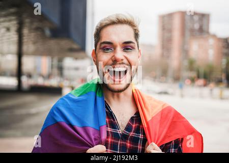 Schwuler Mann mit Make-up auf Spaß tragen lgbt Regenbogenflagge im Freien - Fokus auf Gesicht Stockfoto