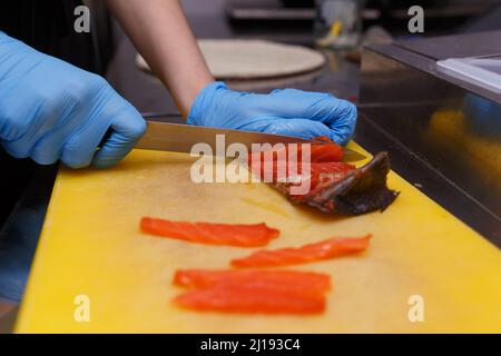 Die Hände des Kochs in hygienischen Handschuhen mit einem scharfen Messer trennen die Lachsfilets von den Knochen. Schneiden von frischem Fisch in der Küche auf einem Schneidegut Stockfoto