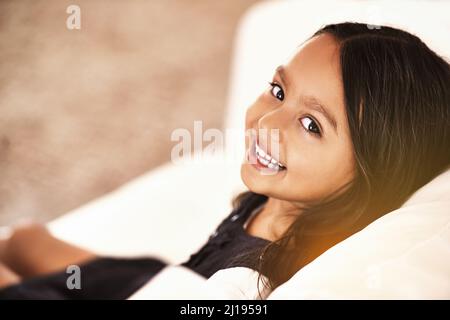 Nur das süßeste Mädchen. Beschnittenes Porträt eines kleinen Mädchens, das zu Hause auf dem Sofa sitzt. Stockfoto