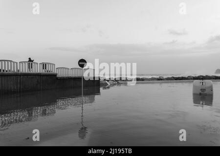 Eine Graustufenaufnahme einer Brücke, einer Steinbarriere und einem Stoppschild, das sich im Wasser spiegelt. Stockfoto