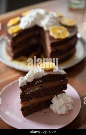 Ein Stück köstlicher veganer Orangenschokoladenkuchen, serviert mit Schlagsahne auf einem Teller mit der ganzen Torte im Hintergrund. Stockfoto