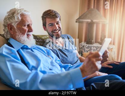 Als Nächstes richten Sie Ihre E-Mail-Adresse ein. Aufnahme eines jungen Mannes, der seinem älteren Großvater zeigt, wie man ein Tablet benutzt, während er zu Hause auf der Couch sitzt. Stockfoto