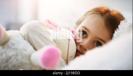 Das süßeste Gesicht der Welt. Aufnahme eines entzückenden kleinen Mädchens, das mit ihrem Teddybären zu Hause im Bett liegt. Stockfoto