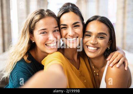 Drei multiethnische junge Freundinnen, die gemeinsam lachen und Selfie im Freien machen Stockfoto