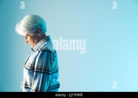 Resignierte, ältere, grauhaarige Großmutter, die ihren Kopf enttäuscht senkte, Seitenansicht, mittellange Studioaufnahme mit blauem Hintergrund. Hochwertige Fotos Stockfoto