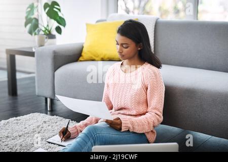 Es gibt nichts auszusetzen an der Arbeit am Wochenende. Ausgeschnittene Aufnahme einer jungen schönen Frau, die Papierkram durchläuft und einen Laptop benutzt, während sie darauf sitzt Stockfoto