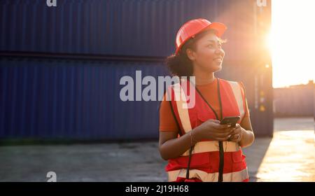 Porträt einer glücklichen, lächelnden afroamerikanischen Ingenieurin, die Sicherheitsweste und Helm trägt, auf logistischer Transportfracht stehend und mit dem Mobiltelefon unterwegs ist Stockfoto