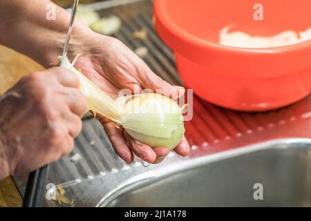 Nahaufnahme einer Köchin in der Küche am Waschbecken, die die Haut mit einem Messer von einer Zwiebel abschält, Deutschland Stockfoto