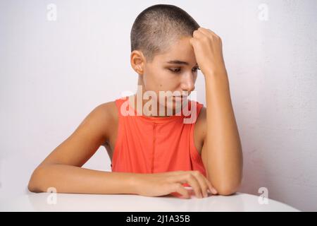 Das kaukasische Mädchen mit kurzen Haaren, fast kahl, hält ihre Hände hinter ihrem Kopf und lehnt sich in einem orangefarbenen T-Shirt auf einem hellen Hintergrund in Nahaufnahme alarmierend auf den Tisch. Das Konzept von Krankheit und Einsamkeit Stockfoto