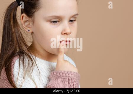 Porträt von jungen niedlichen traurig nachdenklichen blauäugigen Mädchen mit langen dunklen Haaren in weiß und rosa Pullover stehen mit Zeigefinger auf den Mund Stockfoto