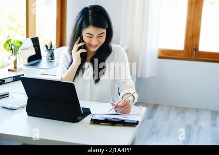 Geschäftsbeziehungen, ist ein asiatischer Sekretär des Unternehmens am Telefon mit Kunden kommt mit Stift in Kontakt, um die Zeit aufzuschreiben, um die zusammenzufassen Stockfoto