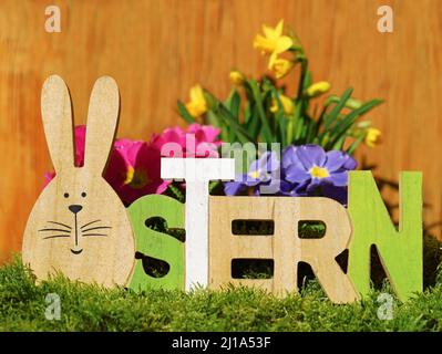 osterhase als O im deutschen Wort, ostern, geschrieben in Holzschrift Decoraction auf Moos vor bunten Frühlingsblumen Stockfoto