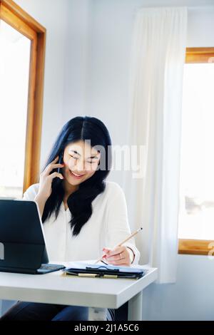 Geschäftsbeziehungen, ist ein asiatischer Sekretär des Unternehmens am Telefon mit Kunden kommt mit Stift in Kontakt, um die Zeit aufzuschreiben, um die zusammenzufassen Stockfoto