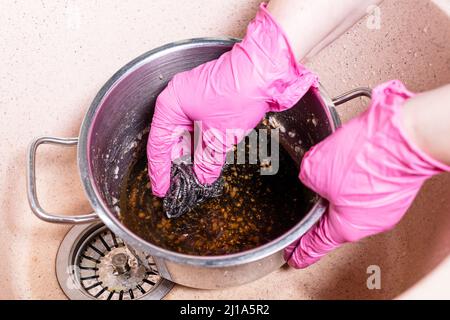 Oben sehen Sie die Handschrubben Stewpan mit verbrannten Lebensmitteln mit Metallschwamm in rosa Waschbecken in der Küche zu Hause Stockfoto