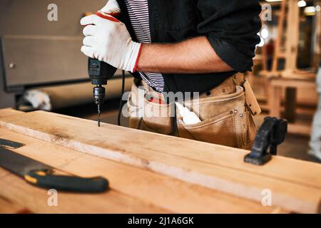 Alles ist vom Bohrer getrennt. Ausgeschnittene Aufnahme eines unerkennbaren Zimmermanns mit einer elektrischen Bohrmaschine in einer Werkstatt. Stockfoto