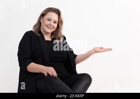 Porträt einer lächelnden fetten, dicken Frau mittleren Alters in schwarzer Strickjacke, Leggings, sitzend und mit flacher Handfläche in der Hand. Stockfoto