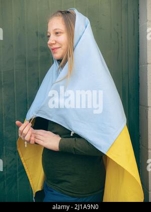 kaukasisch blonde Teenager eingewickelt in einer ukrainischen Flagge über einem neutralen Hintergrund gegen Krieg Konzept Stockfoto