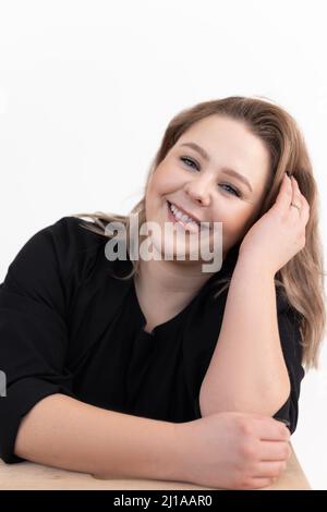 Porträt einer lächelnden, fröhlichen, niedlichen, fetten Frau mittleren Alters mit schwarzer Strickjacke, die am Tisch sitzt und die Zunge zeigt. Stockfoto