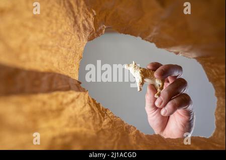 Eine Hand legt eine Figur eines Lammes oder Schafes in einen Papierbeutel. Die Hand eines reifen Mannes hält das Spielzeug eines Kindes über einer offenen braunen Tasche. Von unten nach oben geschossen. Stockfoto